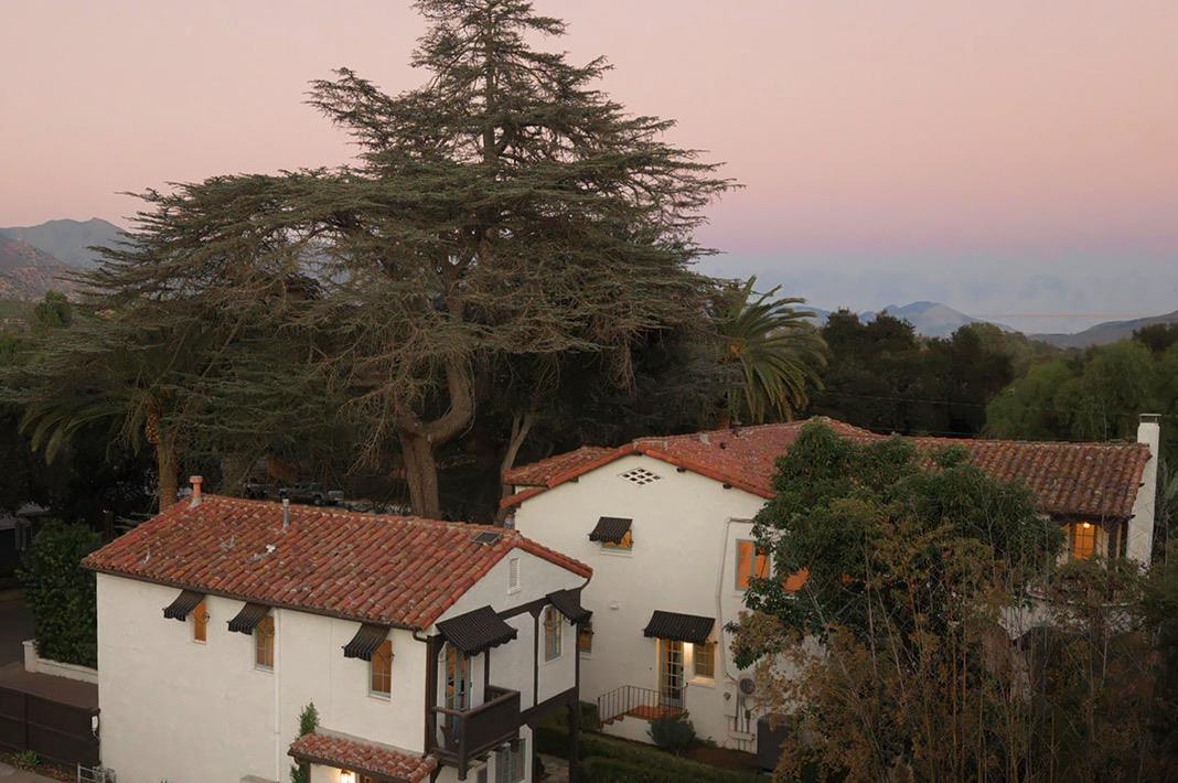 White stucco home with terracotta tile roofs beneath a large cedar tree, with mountains in the distance.