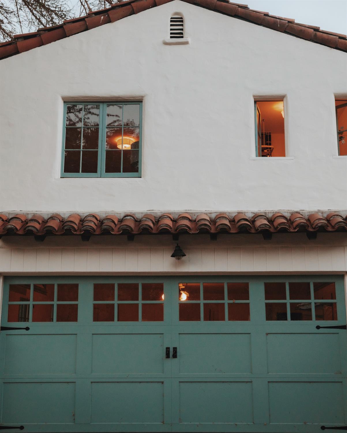 White stucco house with a terracotta tile roof and turquoise garage door, warmly lit at dusk.