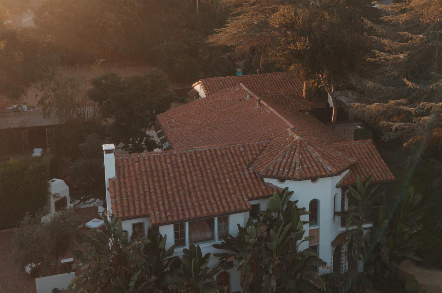 White stucco home with terracotta tile roofs, seen from above among mature trees.
