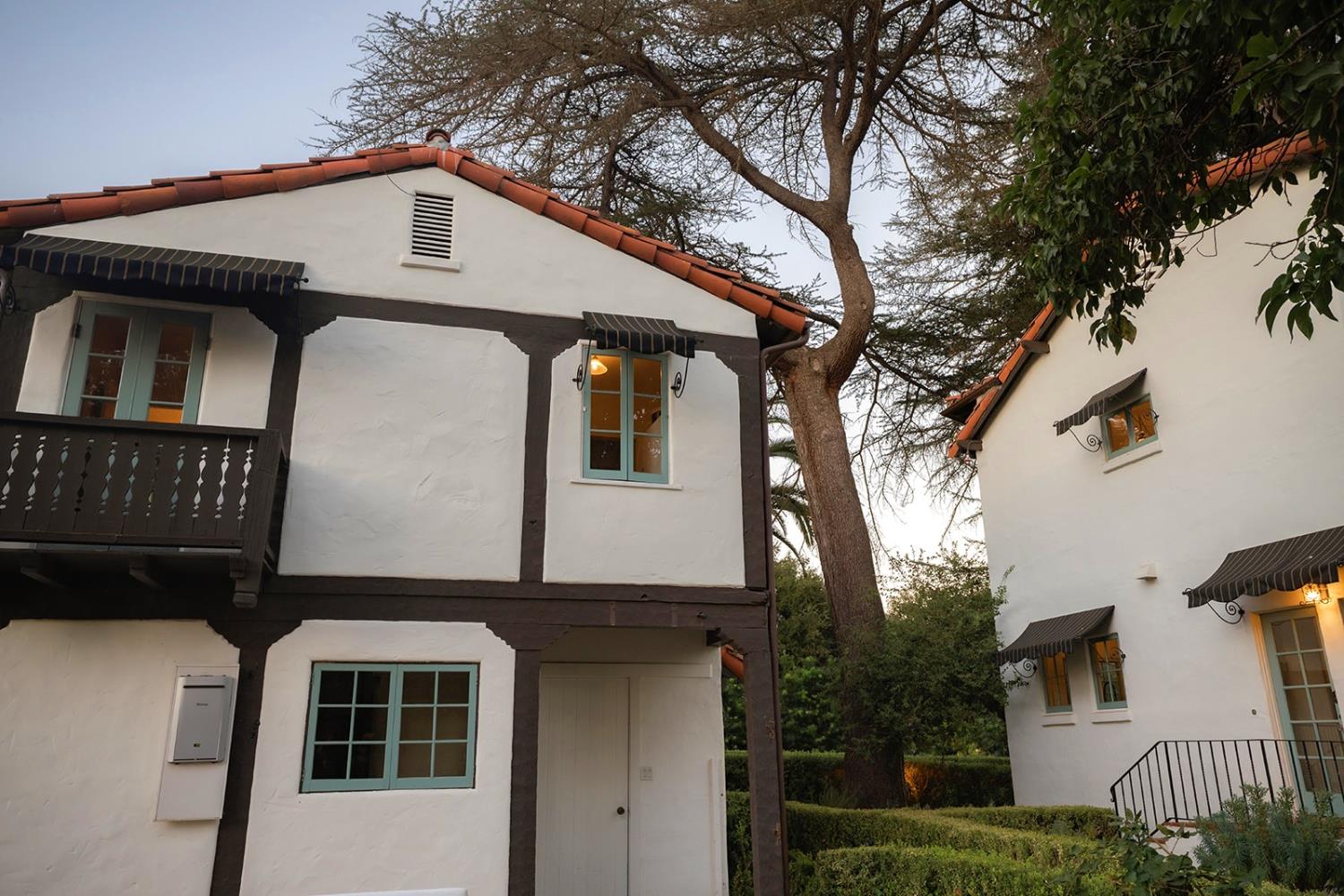 White stucco homes with red tile roofs and teal windows frame a mature tree and clipped hedges.