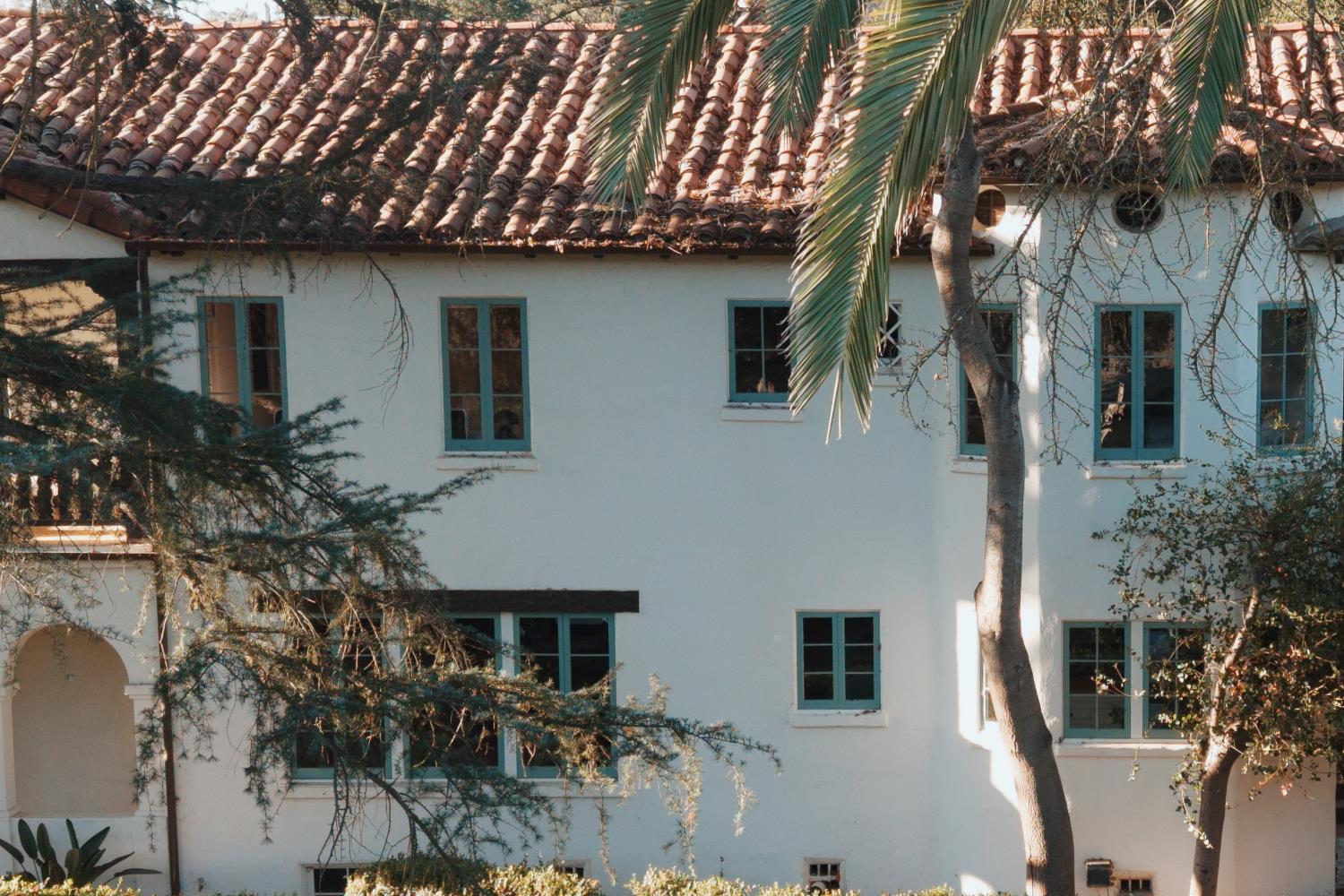 White stucco home with a clay tile roof and teal-trimmed windows, partly screened by trees.
