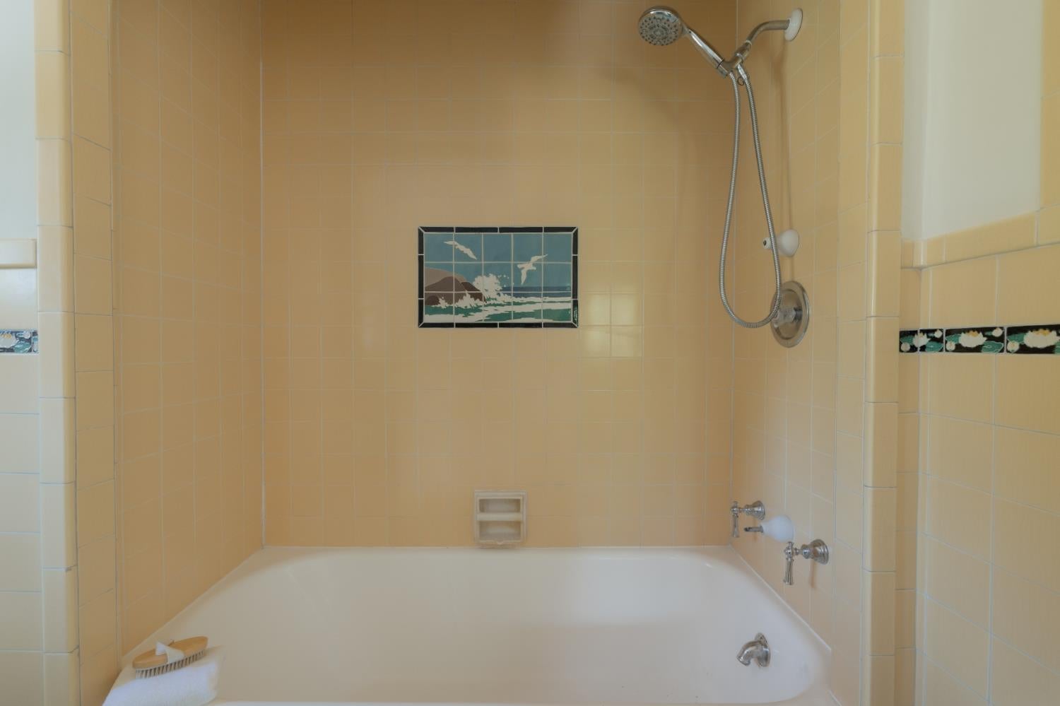 Pale yellow tiled bathtub and shower with a handheld showerhead and ocean-wave tile panel.