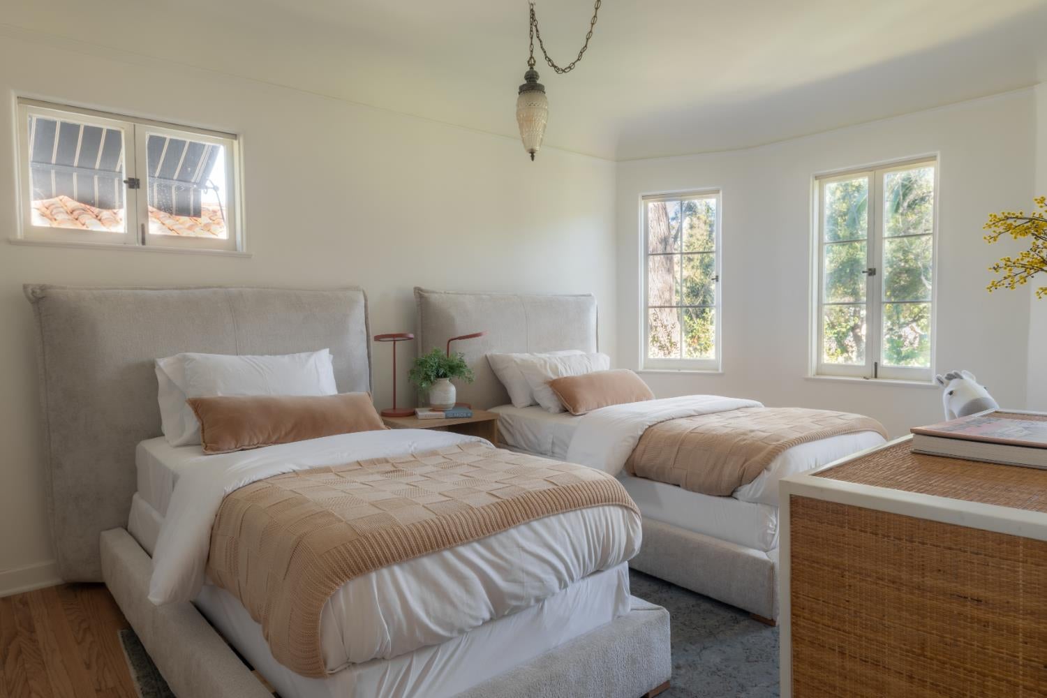 Bedroom with two beds, tan throws, large windows, and a woven-front dresser.