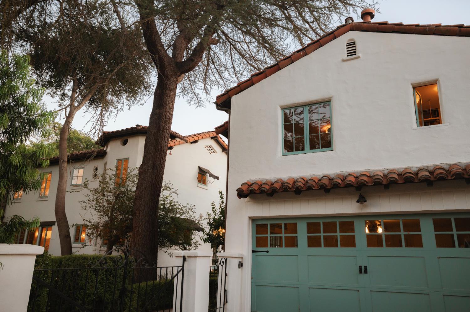 White stucco home with terracotta tile roofs and turquoise garage doors, framed by mature trees.