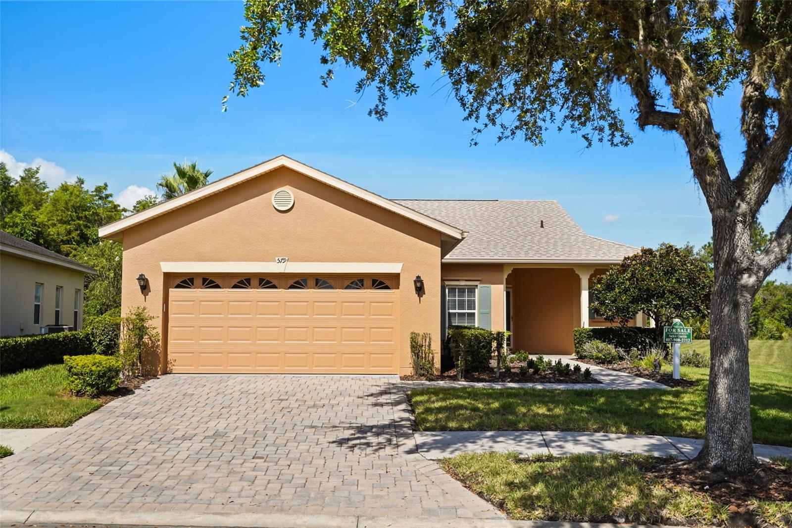 Tan stucco home with a two-car garage, paved driveway, and large shade tree.