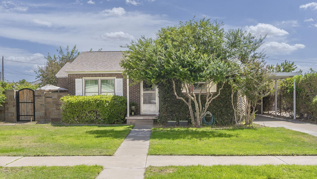 Single-story brick house with a front walkway, mature trees, lawn, stone gate, and covered carport.