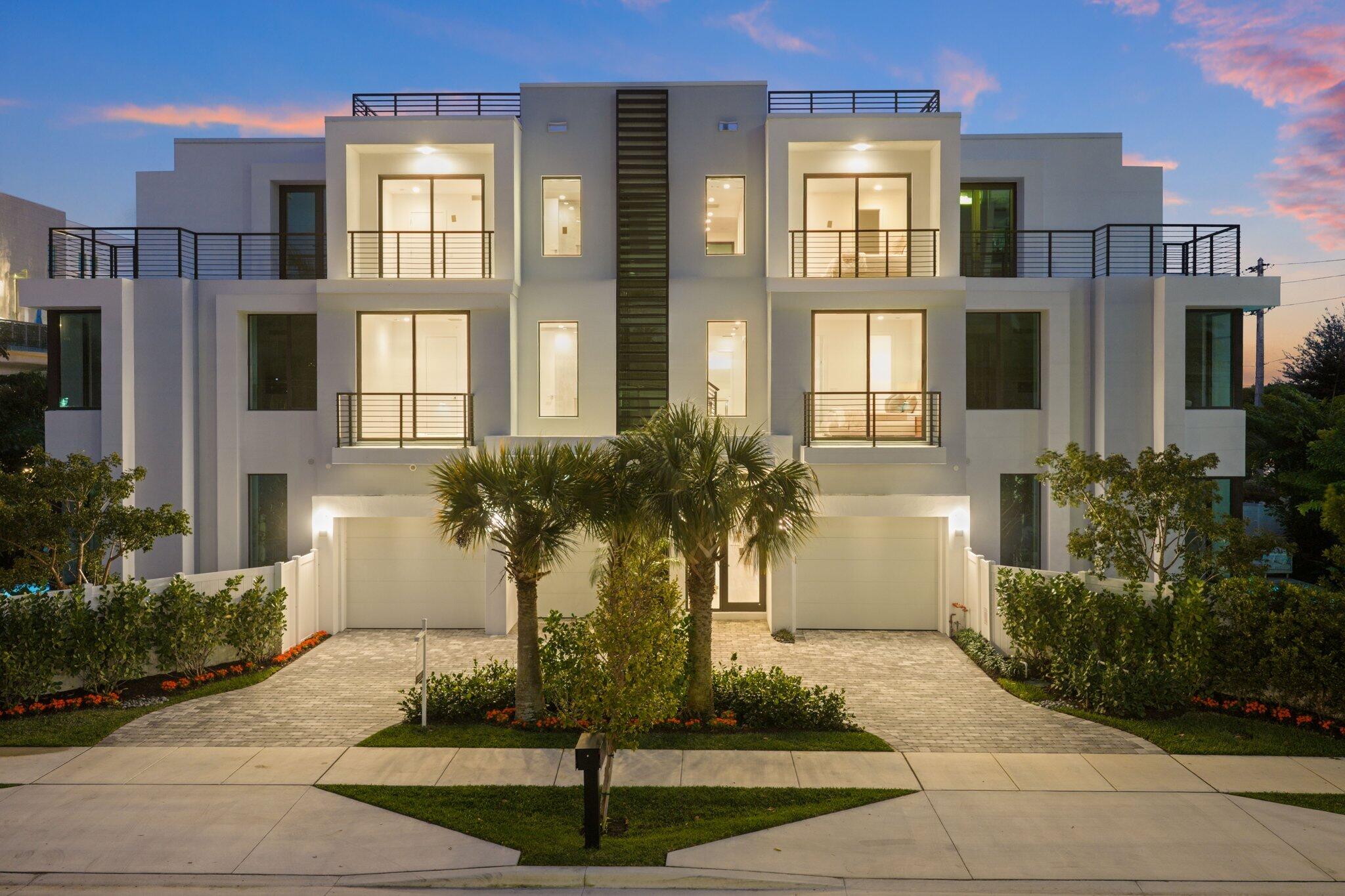 Modern white multi-story home with balconies, palm trees, and illuminated windows at dusk.