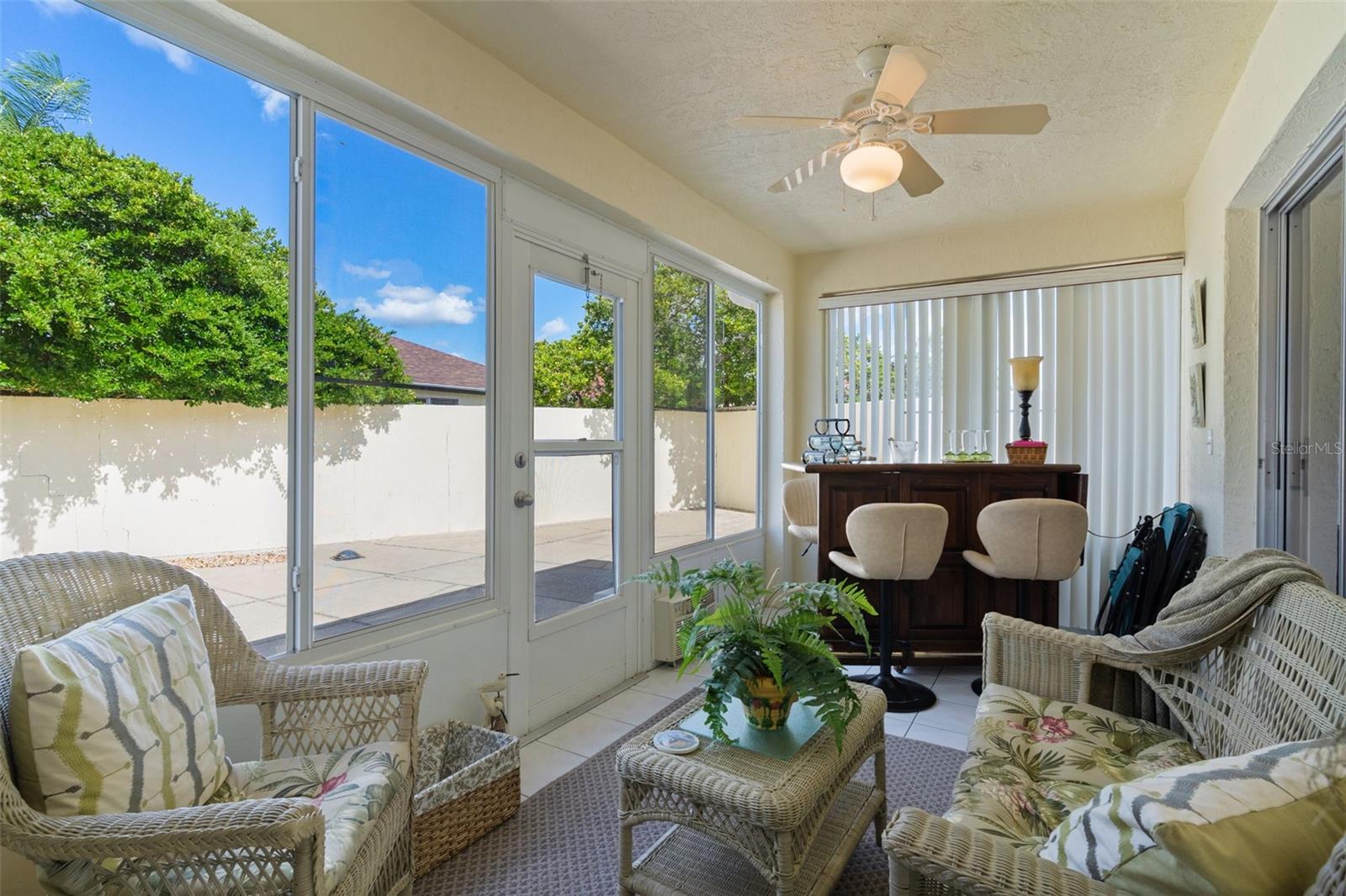 Furnished sunroom with wicker seating and a bar, lined with large windows overlooking a patio.