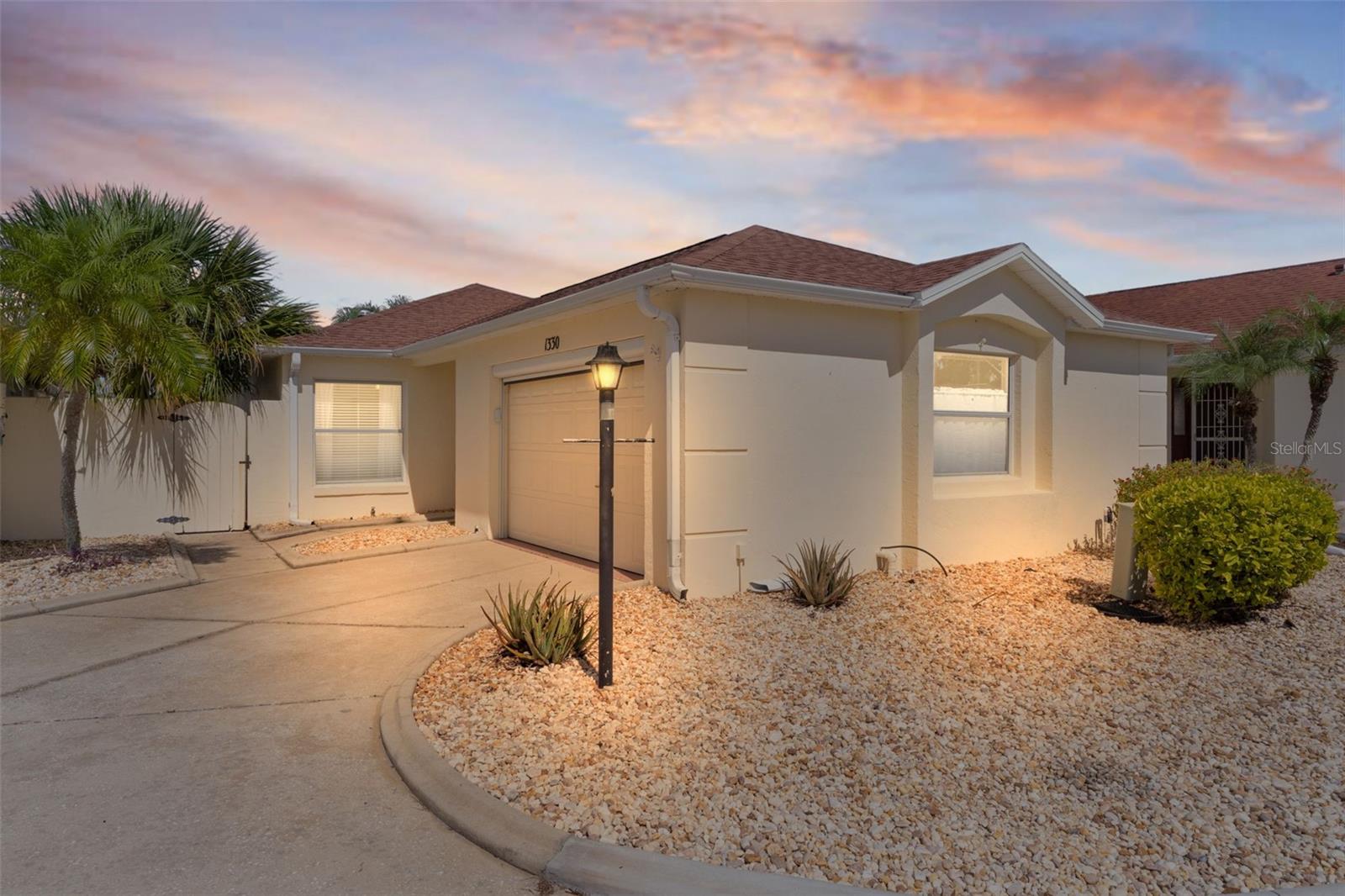 Single-story beige home with a garage, curved driveway, rock landscaping, and palm trees at dusk.