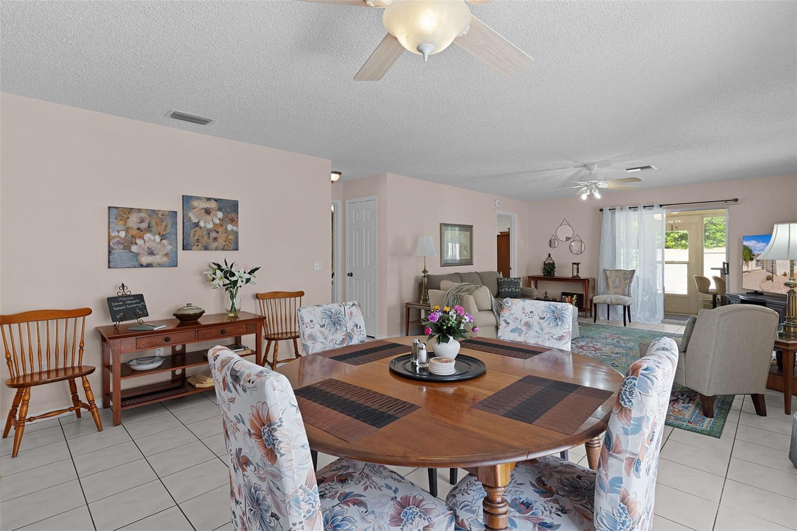 Dining area with a round wooden table, floral chairs, and sliding glass doors overlooking greenery.
