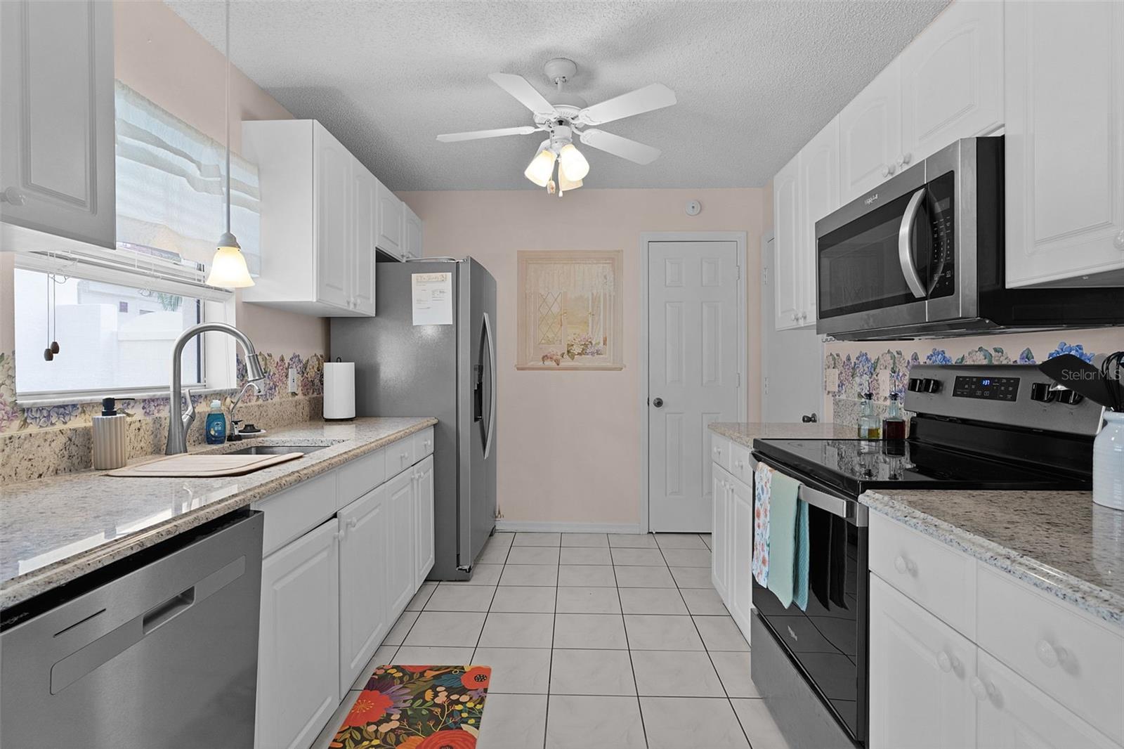 Galley kitchen with white cabinets, stone counters, stainless appliances, and a sink beneath the window.