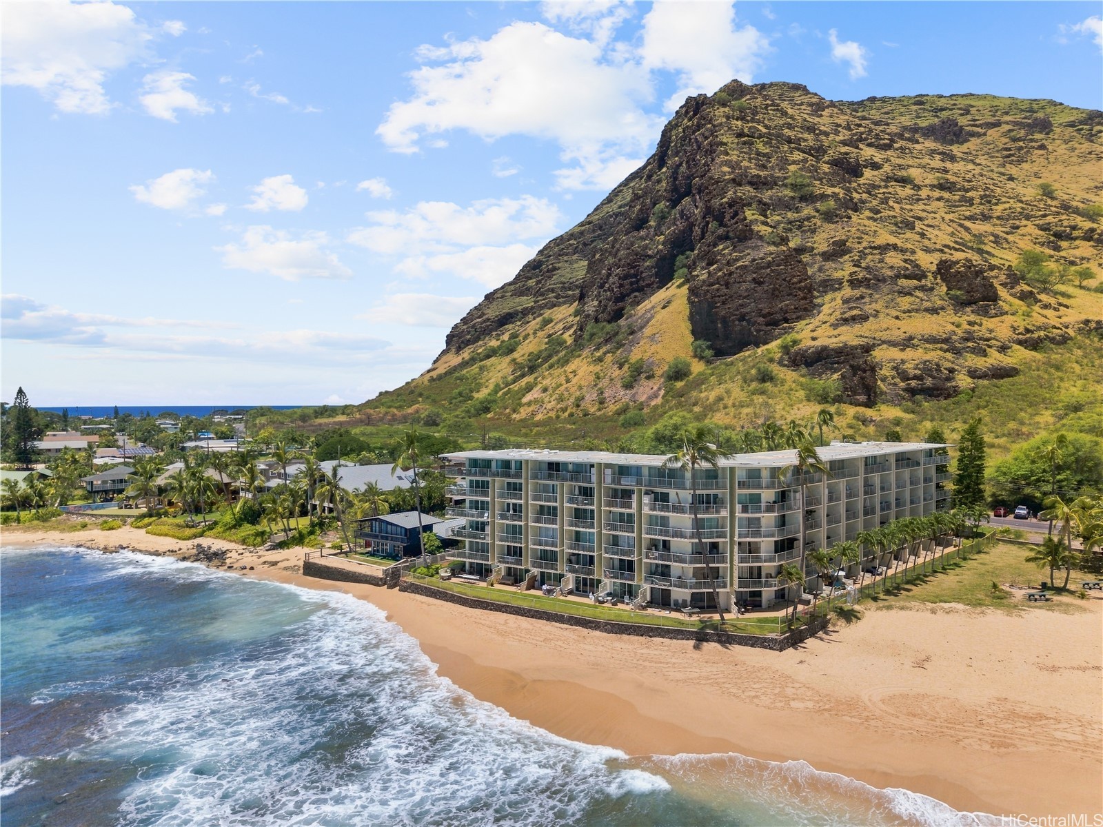 Beachfront condominium beneath a rugged mountain, with ocean waves washing onto the sandy shore.