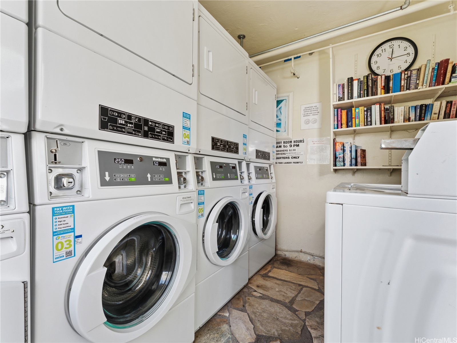 Shared laundry room with front-loading machines, book shelves, and a stone floor.
