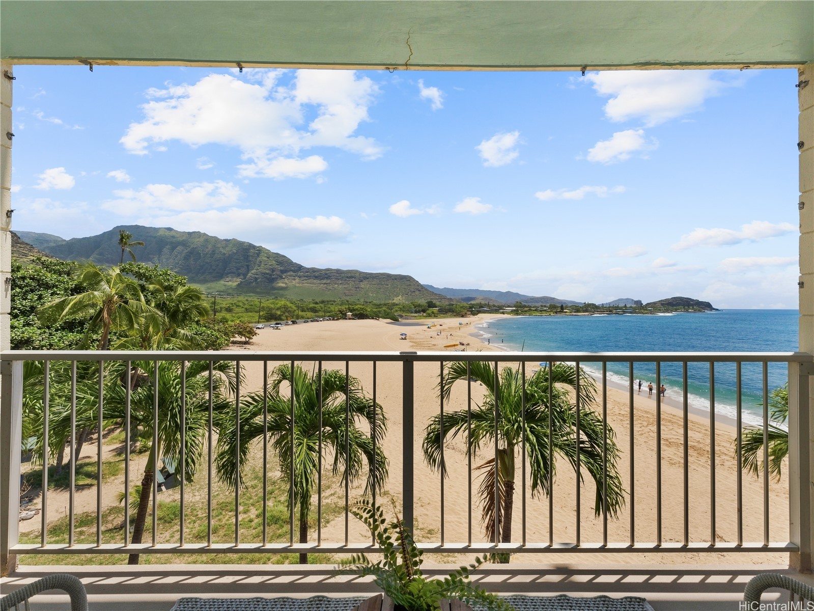 Balcony overlooking a sandy beach and turquoise ocean, framed by palm trees and green mountains.