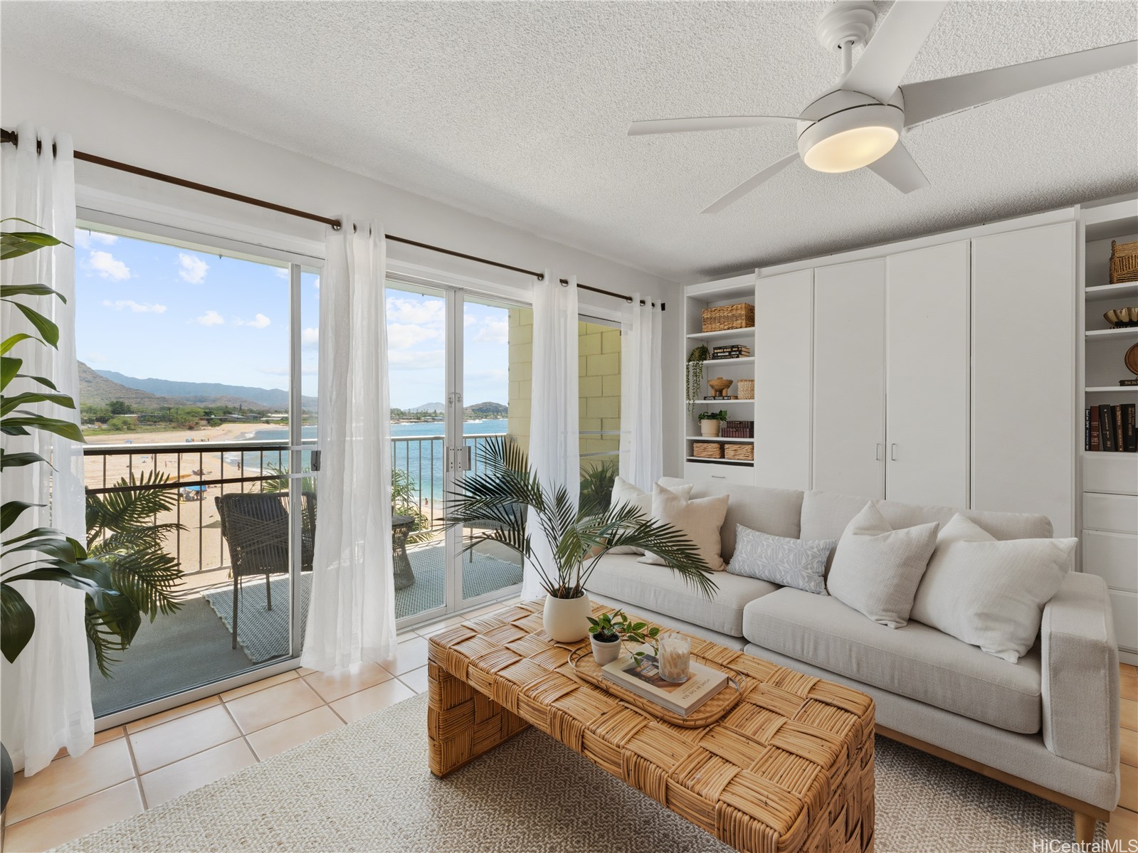Living room with a white sofa and woven coffee table, opening onto a balcony overlooking the beach and ocean.