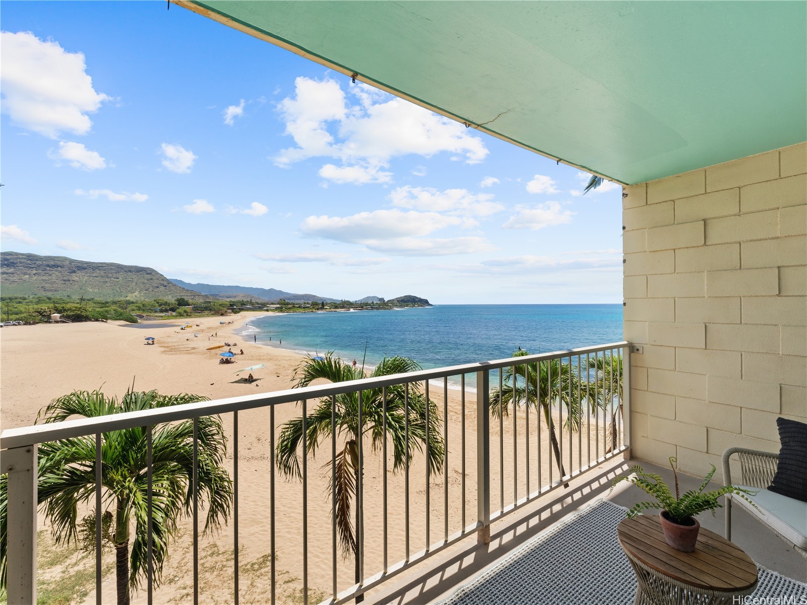 Furnished balcony overlooking a sandy beach and turquoise ocean, with green hills along the coast.
