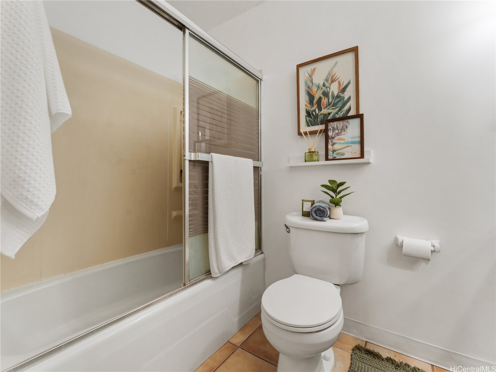 Bathroom with a glass-door bathtub and shower, white toilet, and framed tropical artwork.
