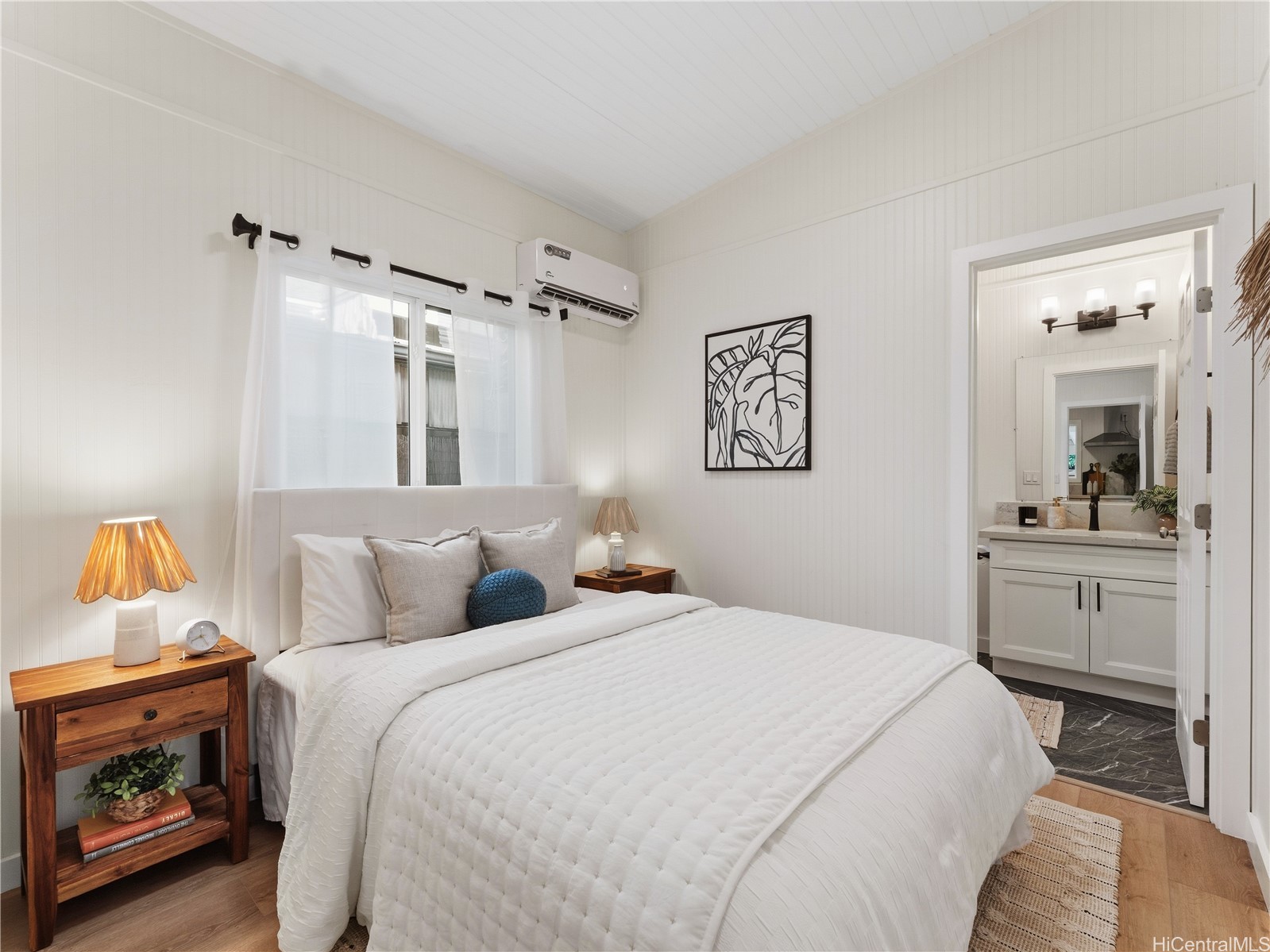 Staged bedroom with a white bed, wood nightstands, paneled walls, and an adjoining bathroom vanity.