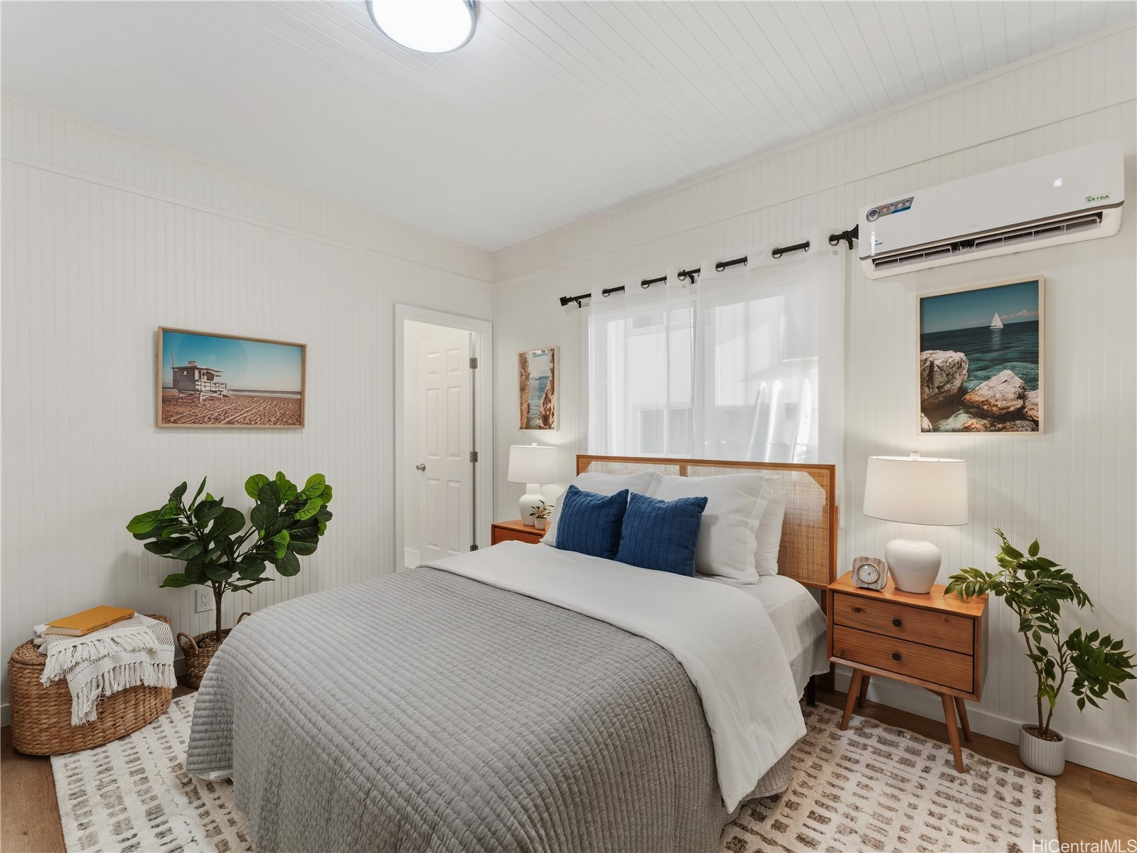Staged bedroom with a large bed, blue accent pillows, white paneled walls, and wall-mounted air conditioner.