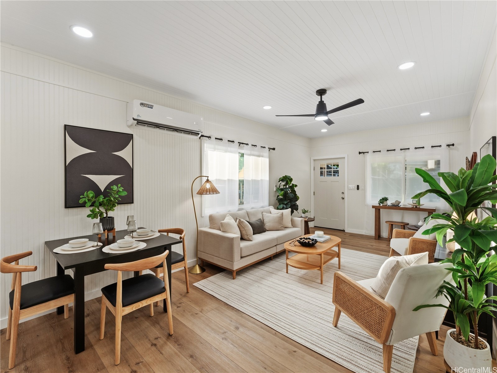 Open-plan living and dining room with a beige sofa, wood floors, and leafy potted plants.