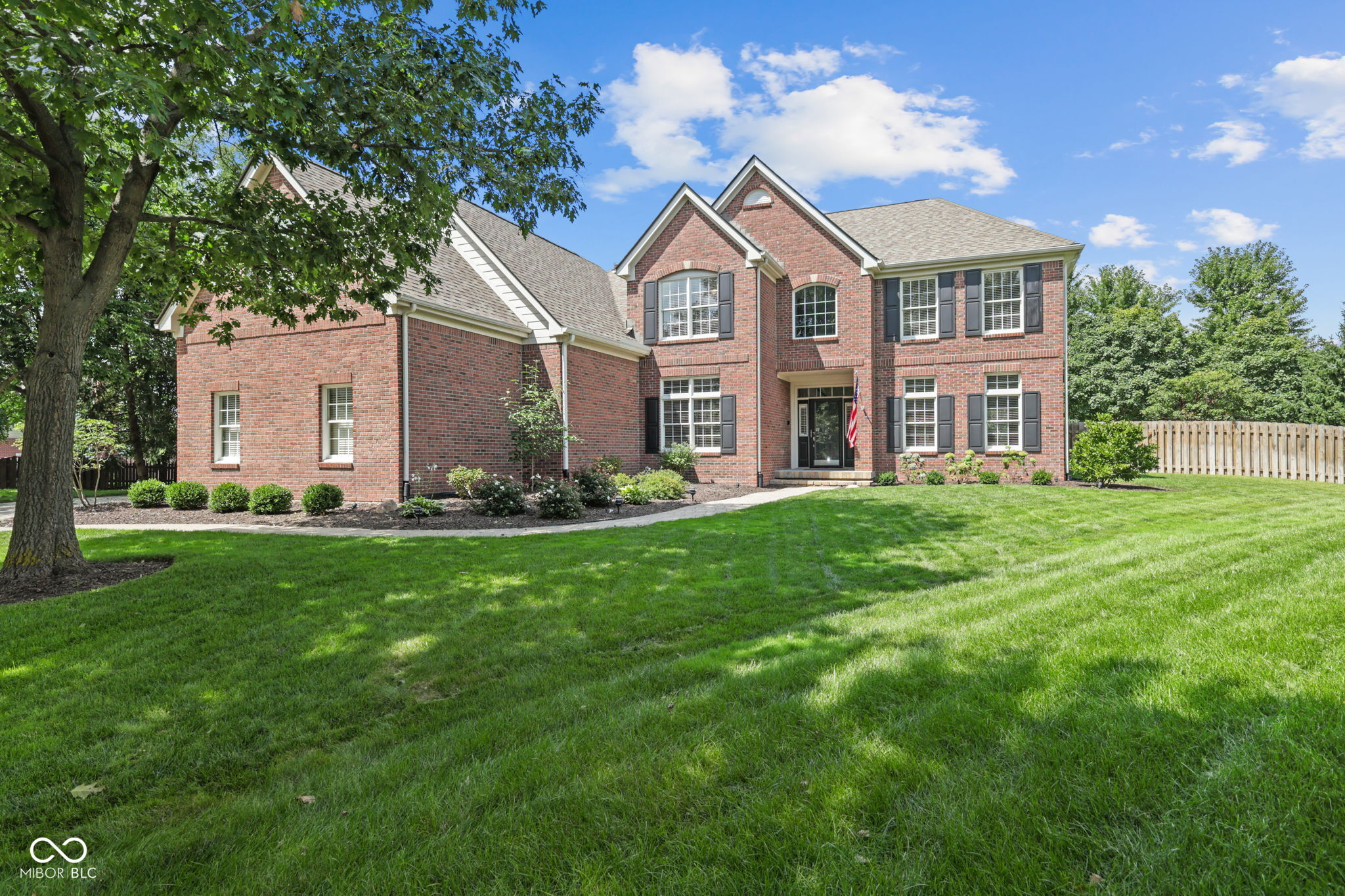 Two-story red brick house with dark shutters, a broad lawn, and a mature shade tree.
