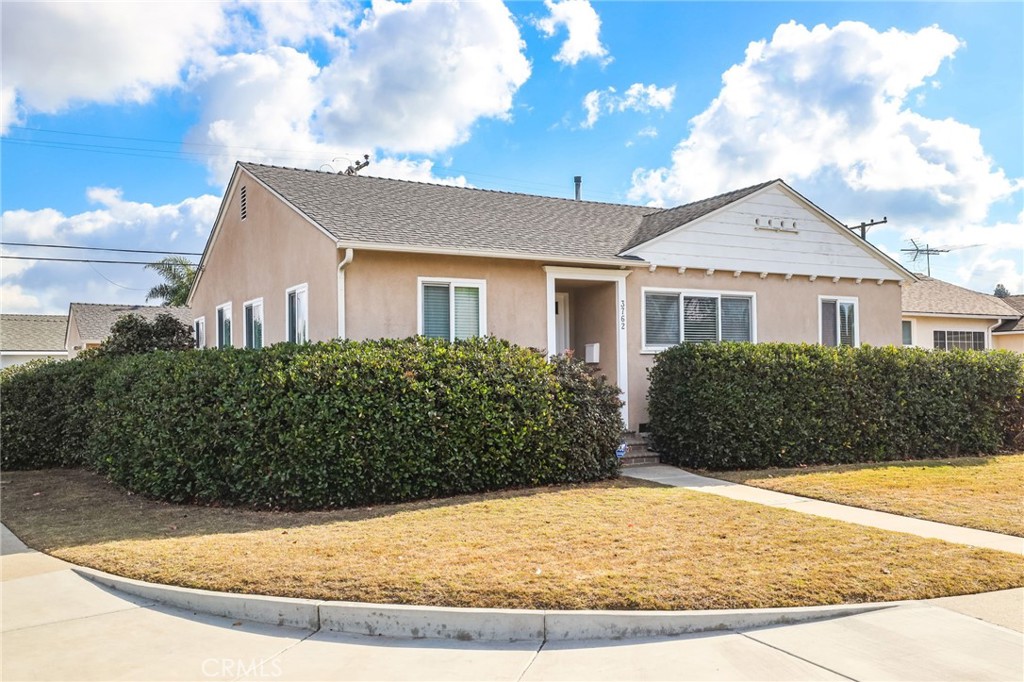 Single-story tan stucco home with a white front gable and a tall hedge along the corner lot.