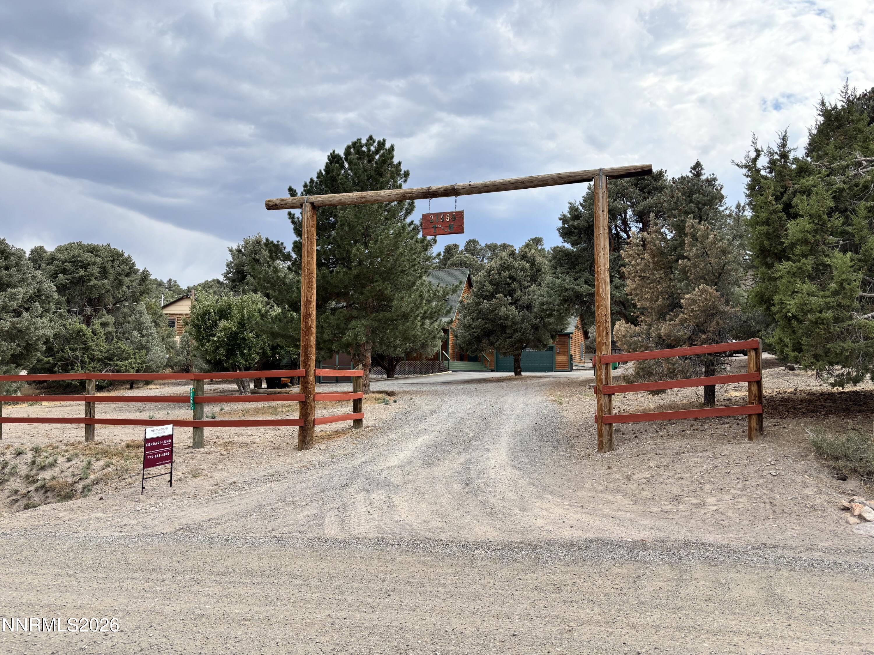 Wooden entrance gate and gravel driveway leading toward a cabin-style house among pine trees.