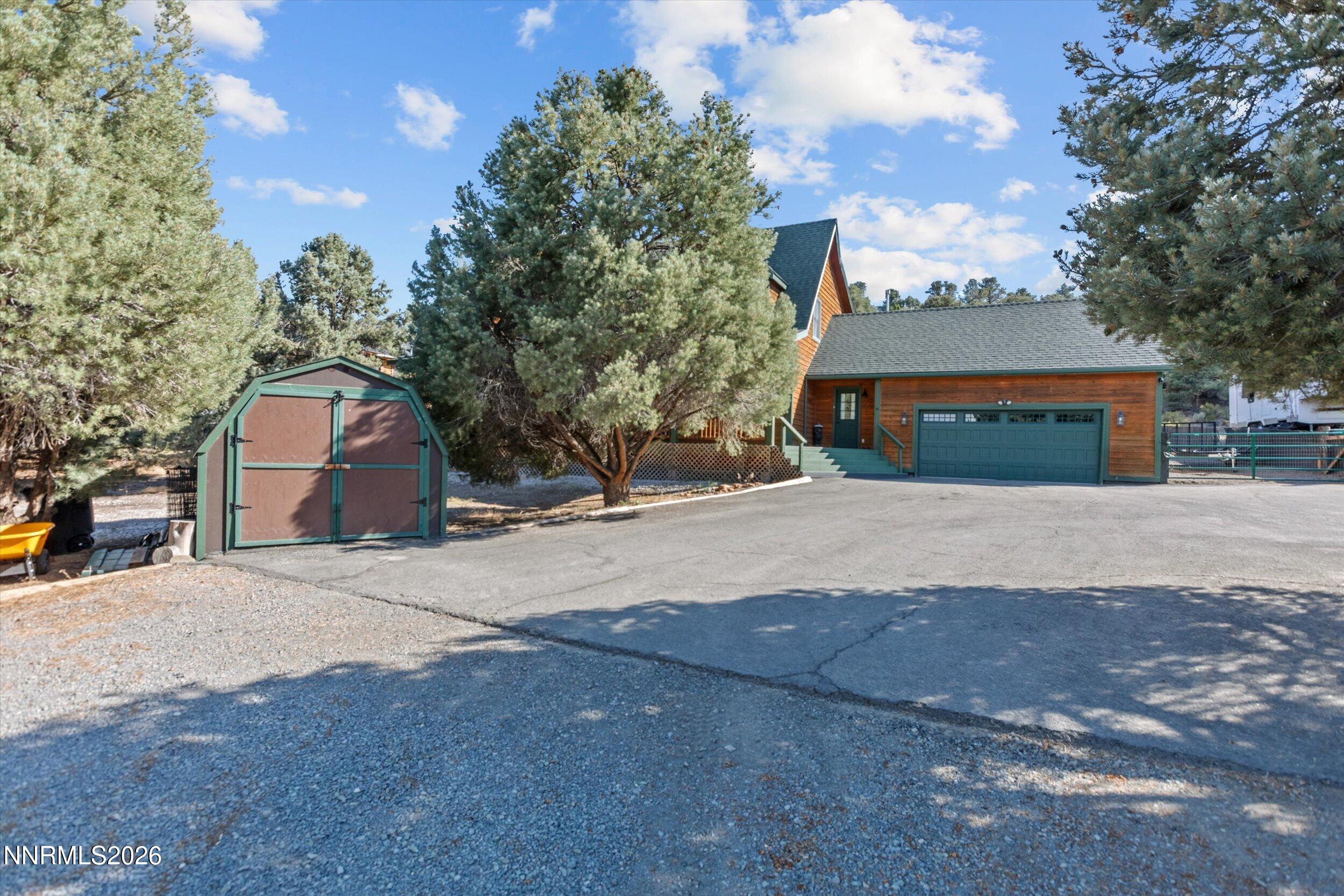 Wood-sided home with a green garage door, detached shed, and wide driveway among pine trees.