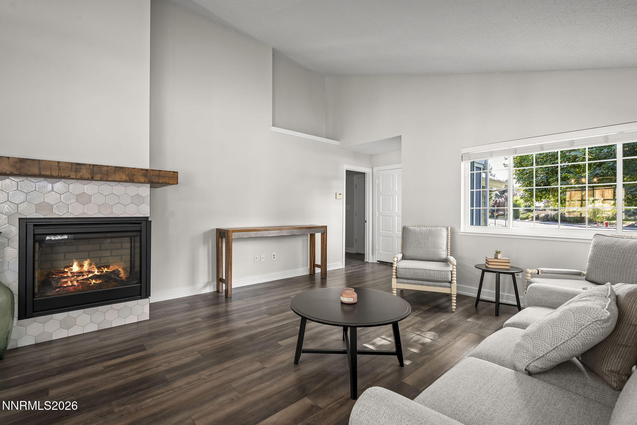 Living room with a wood-mantel fireplace, gray seating, dark floors, and a large window.