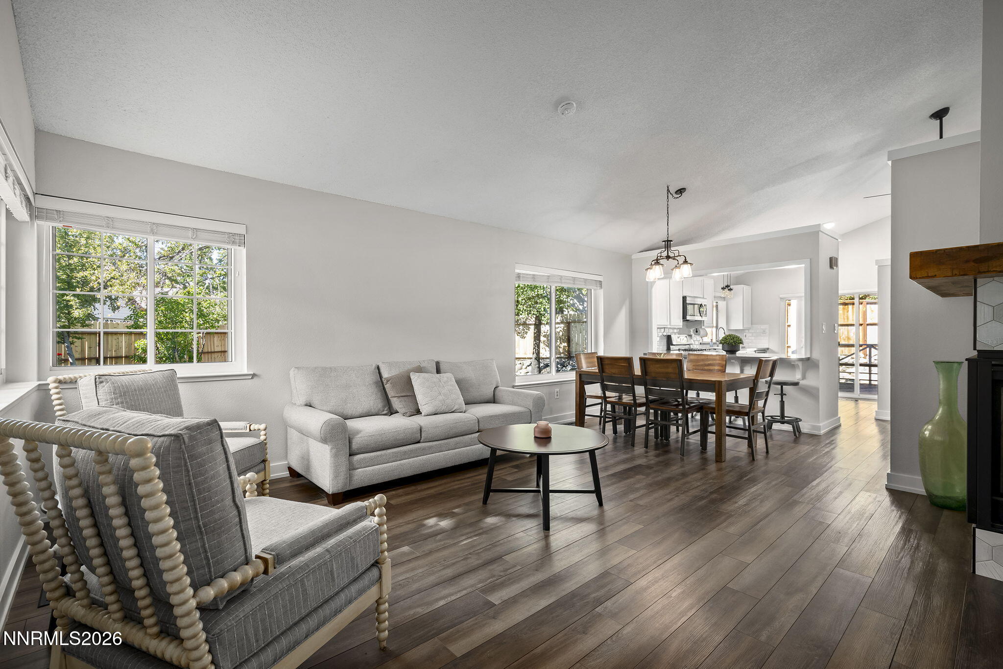 Open-plan living and dining room with gray seating, dark plank floors, and large windows overlooking trees.