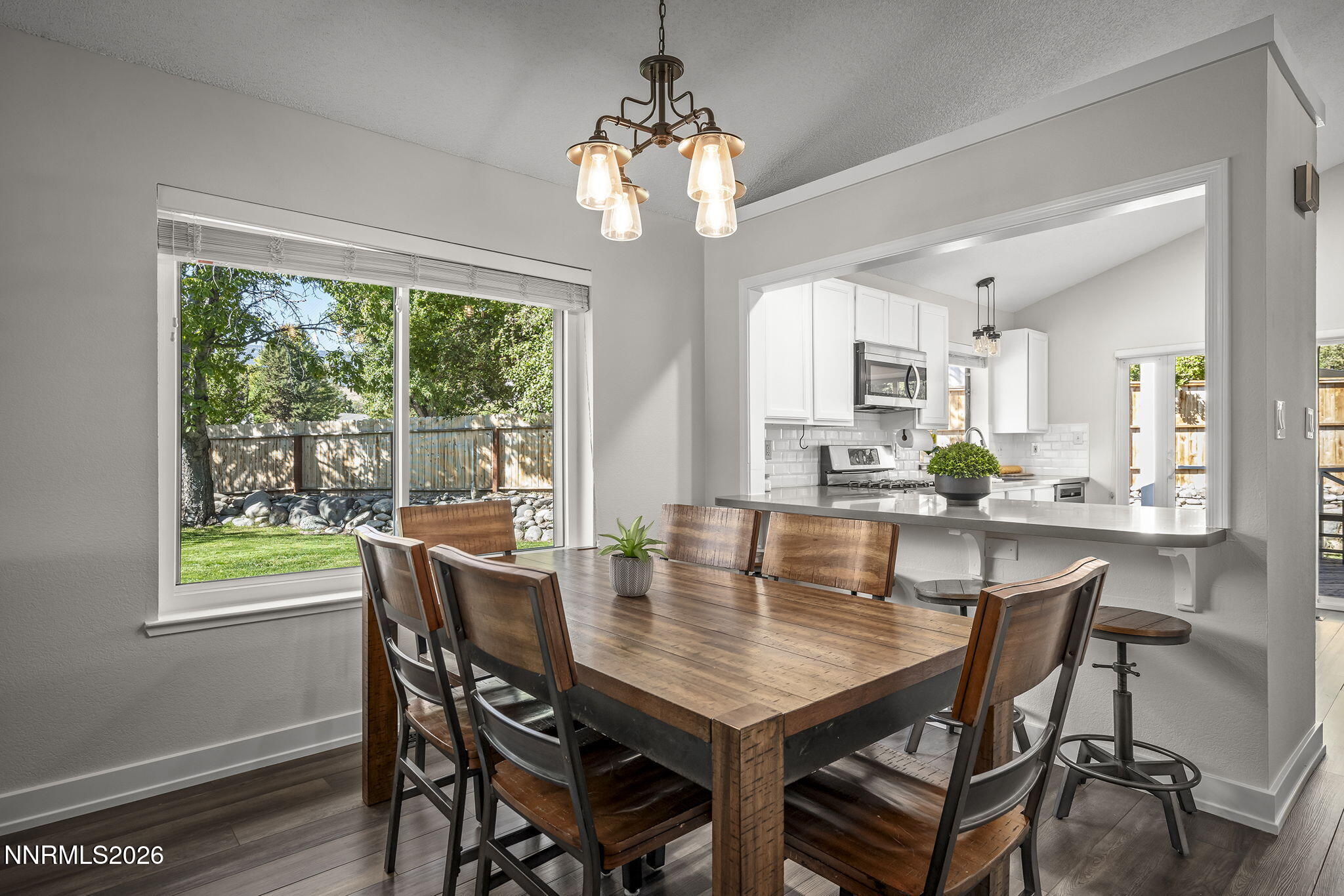 Dining area with a rustic wood table beside a large window, opening to a white kitchen.