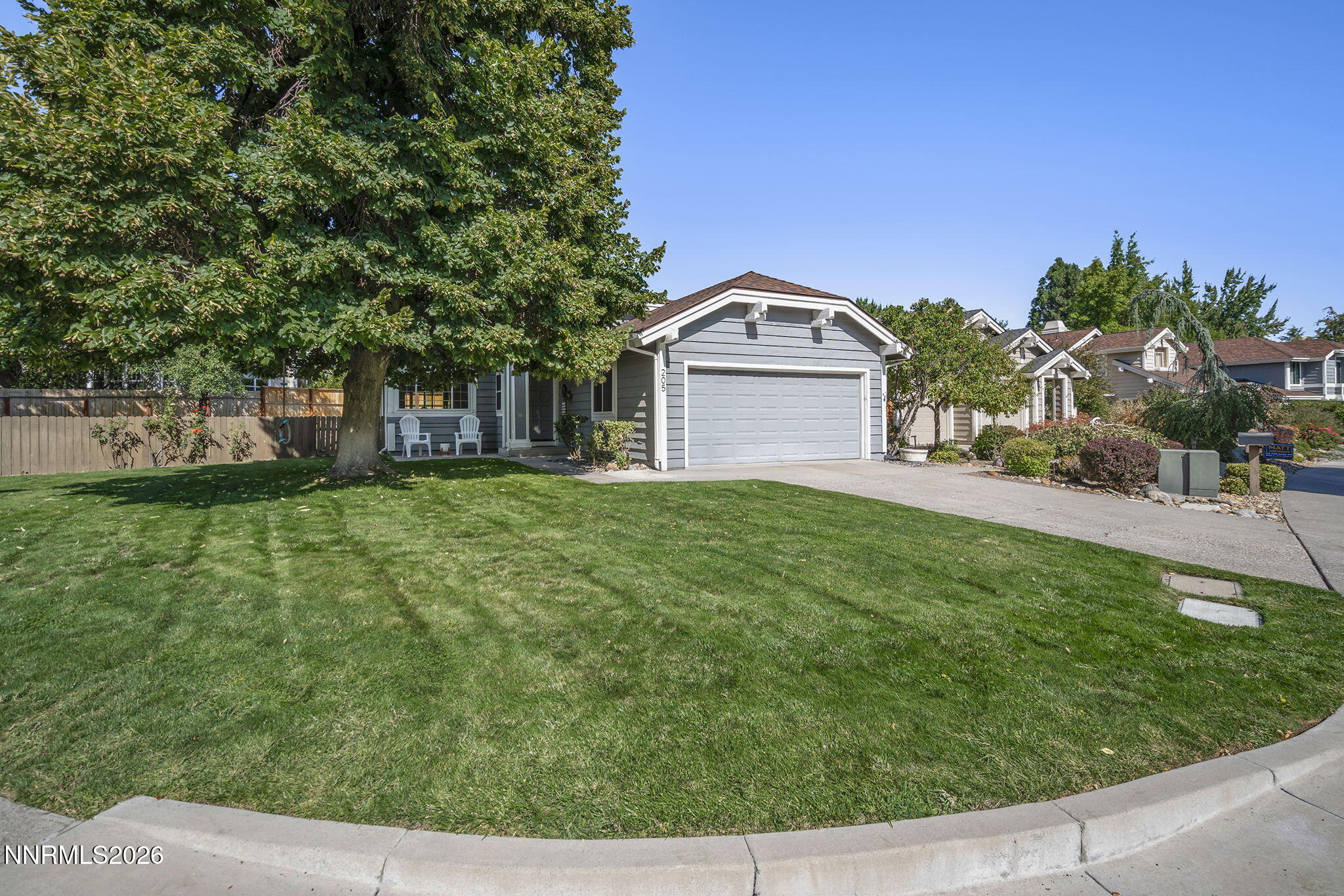 Gray-sided house with a white two-car garage, broad green lawn, mature shade tree, and covered porch.