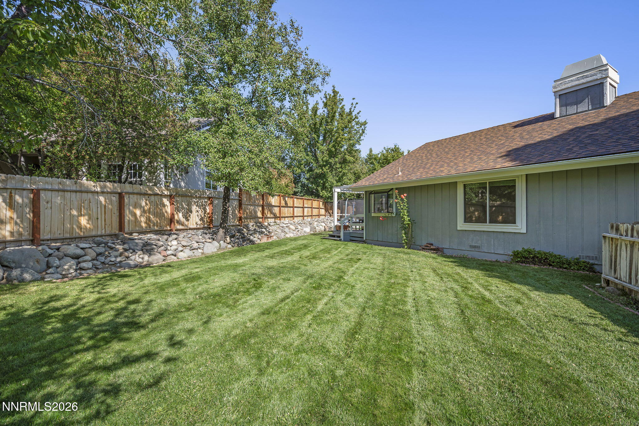 Backyard with a broad lawn, mature trees, rock border, wooden fence, and gray-sided house.