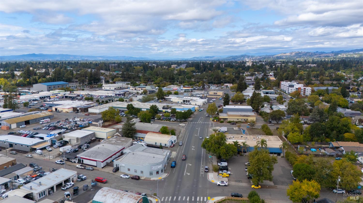 Aerial view of commercial buildings and a major intersection, with tree-lined neighborhoods and hills beyond.