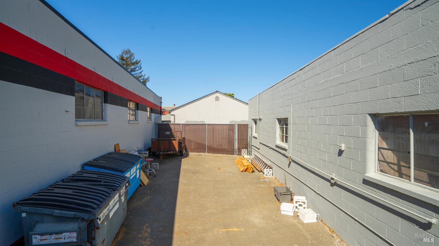 Paved service yard between gray commercial buildings, with dumpsters and a metal gate.