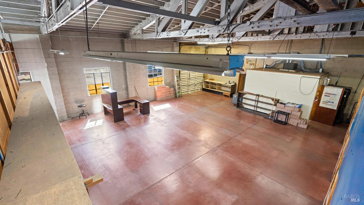 Large workshop interior with exposed roof trusses, fluorescent lights, and a wide reddish floor.