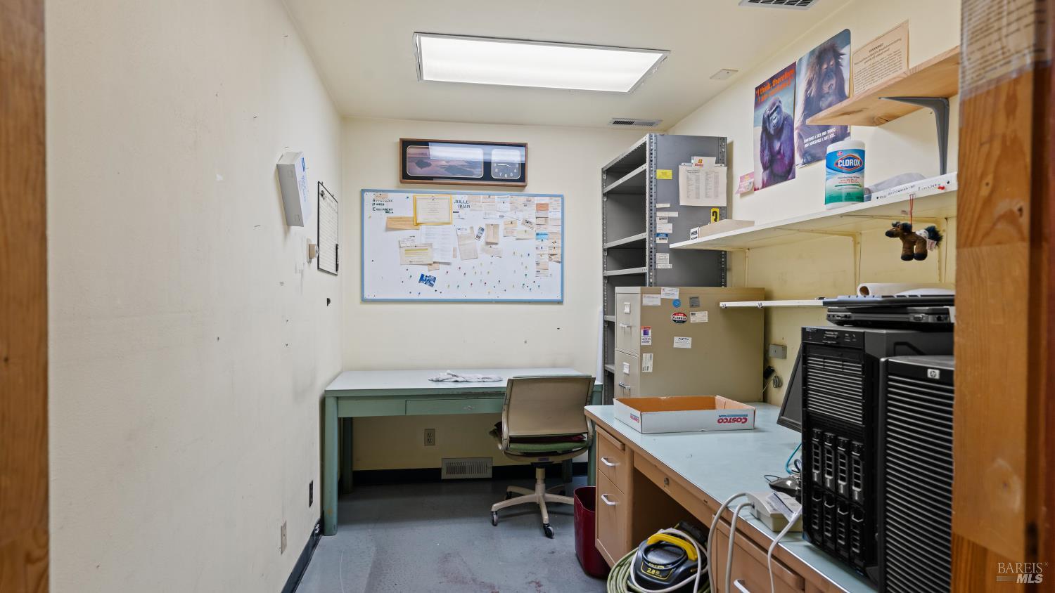 Office workroom with computer equipment, desks, metal shelving, and a bulletin board.