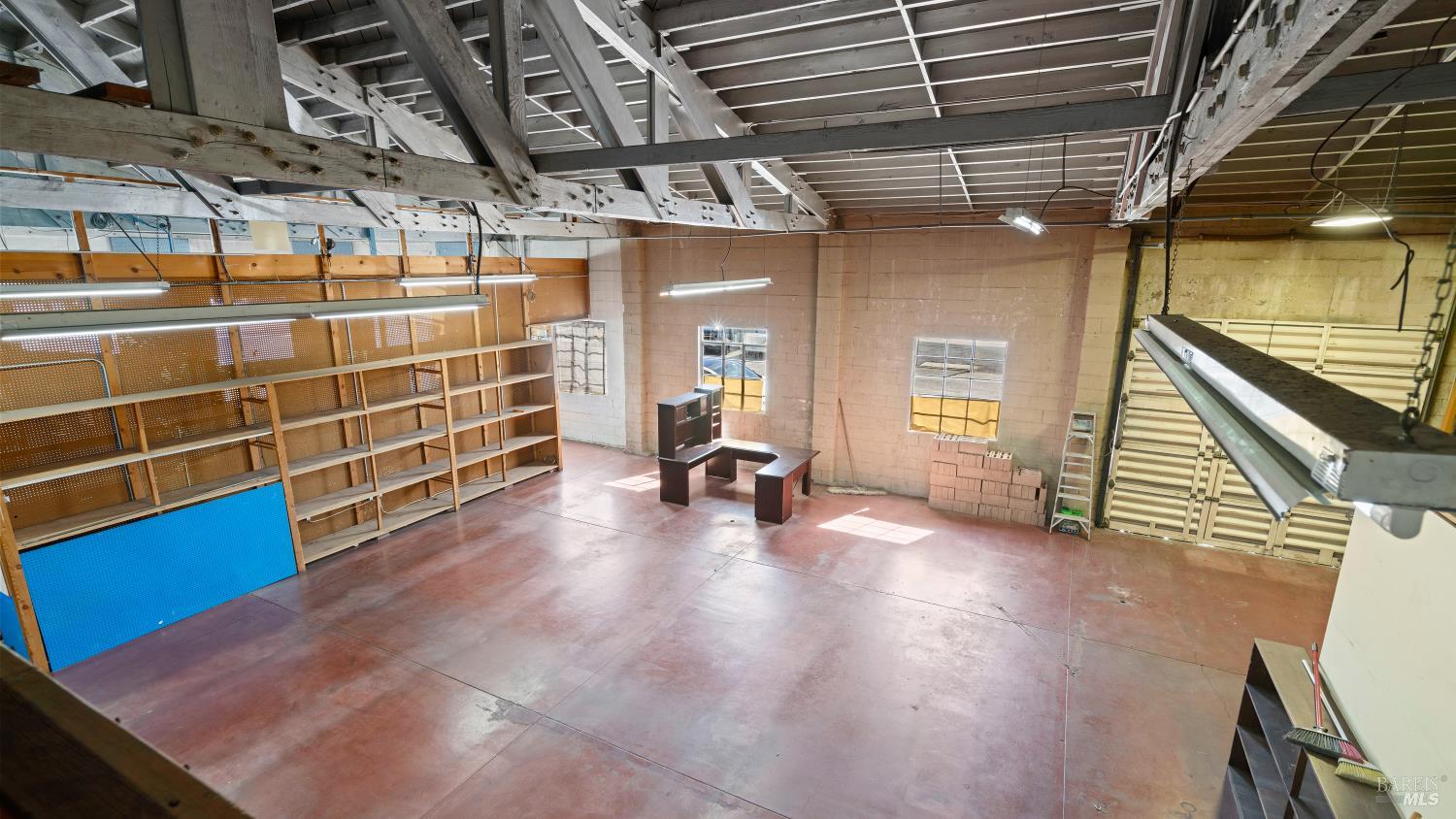 Commercial interior with exposed roof trusses, long shelving, a small desk, and a roll-up door.