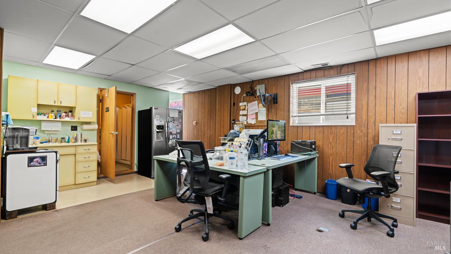 Office interior with green desks and computers, black chairs, wood-paneled walls, and a kitchenette.