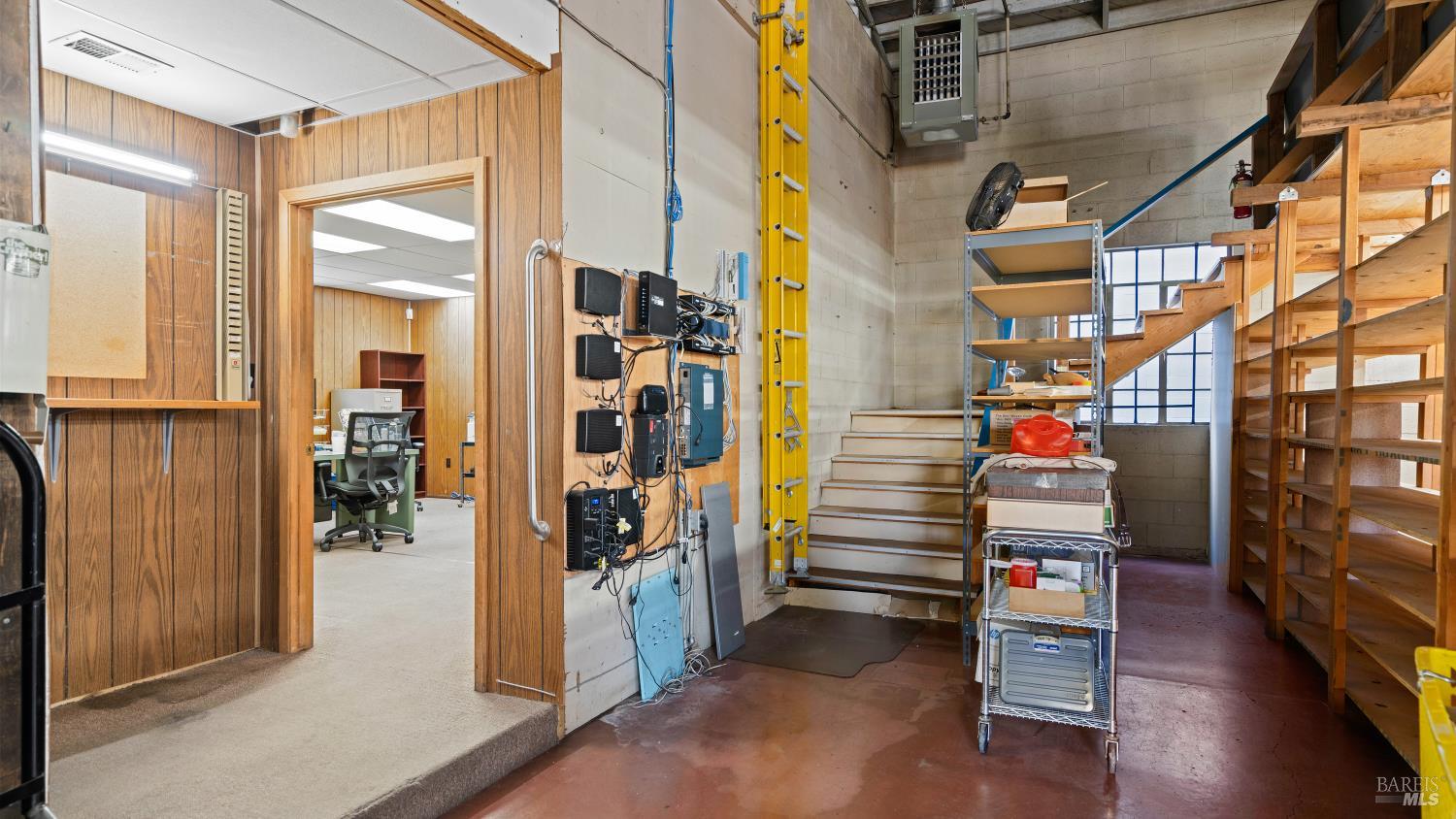 Utility and storage area with wall-mounted equipment, metal shelving, and stairs beside an office doorway.