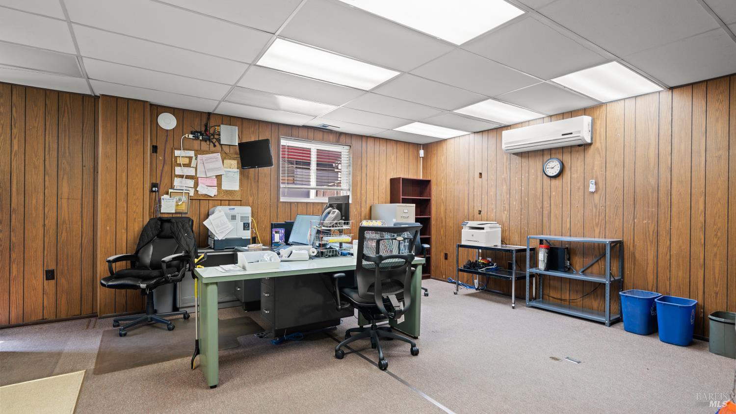 Office with wood-paneled walls, workstation, metal shelving, and wall-mounted air conditioner.
