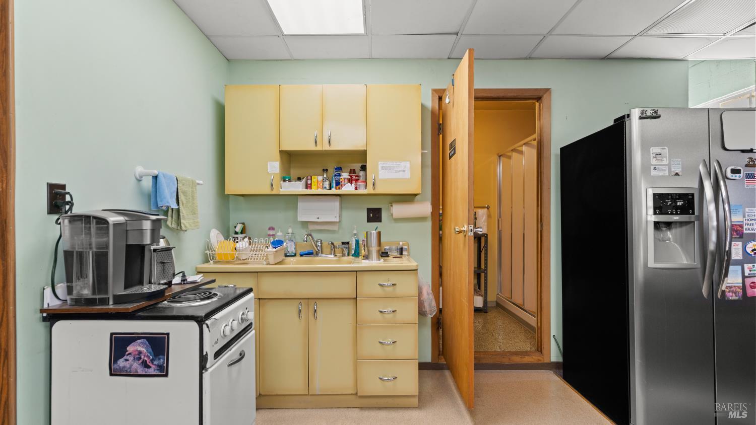 Compact kitchen with pale yellow cabinets, a freestanding stove, and a large stainless-steel refrigerator.