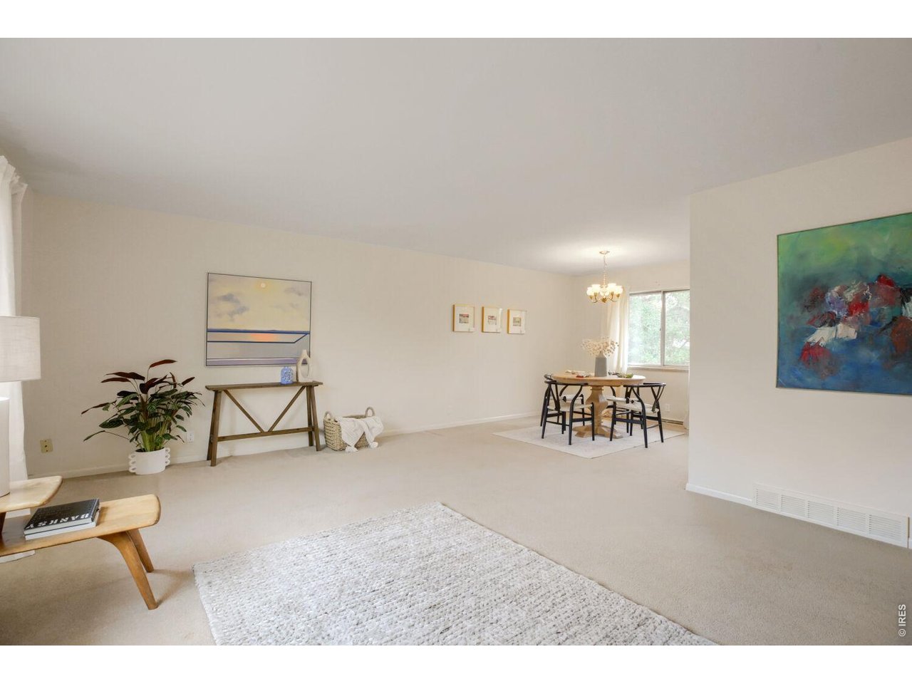Living room opening into a dining area, with neutral carpeting, framed art, and a chandelier.