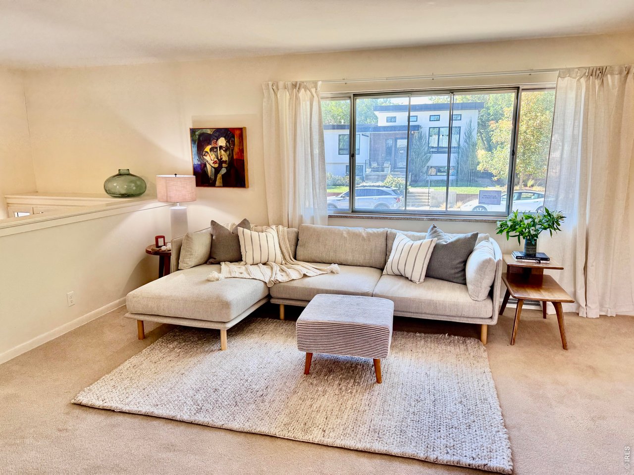 Living room with a beige sectional, striped ottoman, and large window overlooking a modern home.