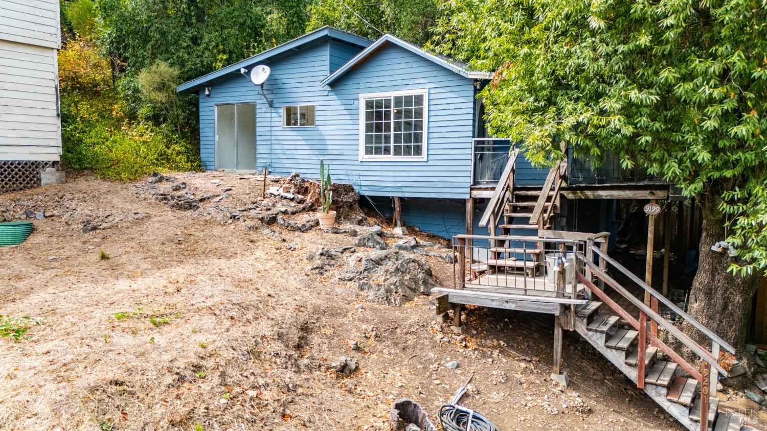 Blue raised house with a wooden deck and stairs on a rocky, tree-lined slope.
