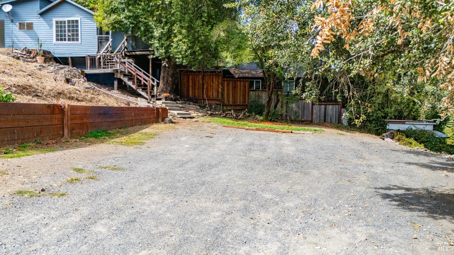 Blue house with a raised deck and stairs above a broad gravel driveway.