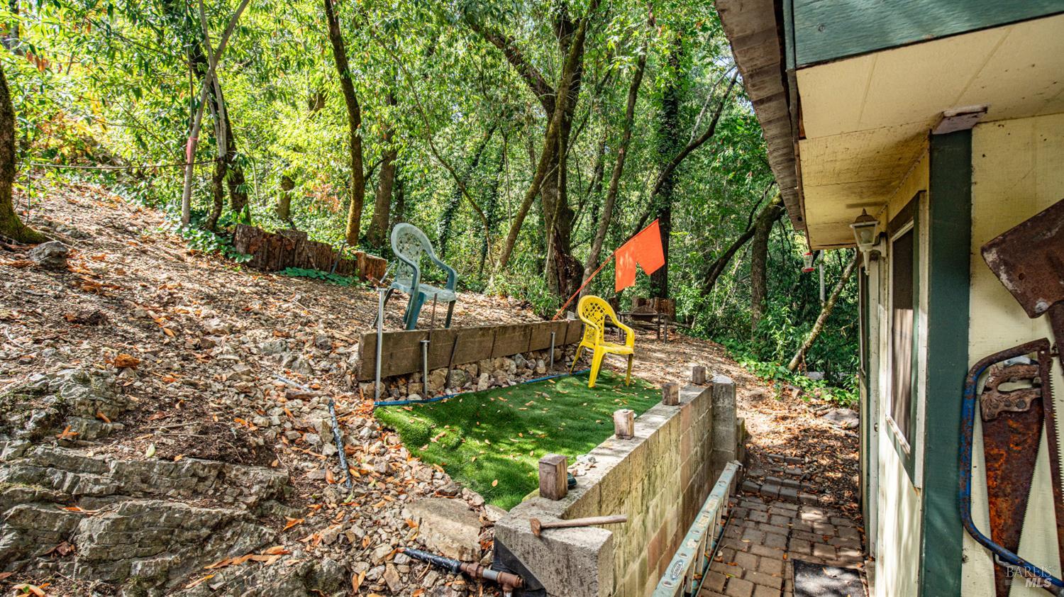 Wooded hillside yard beside a house, with a small turf seating area and stone retaining walls.
