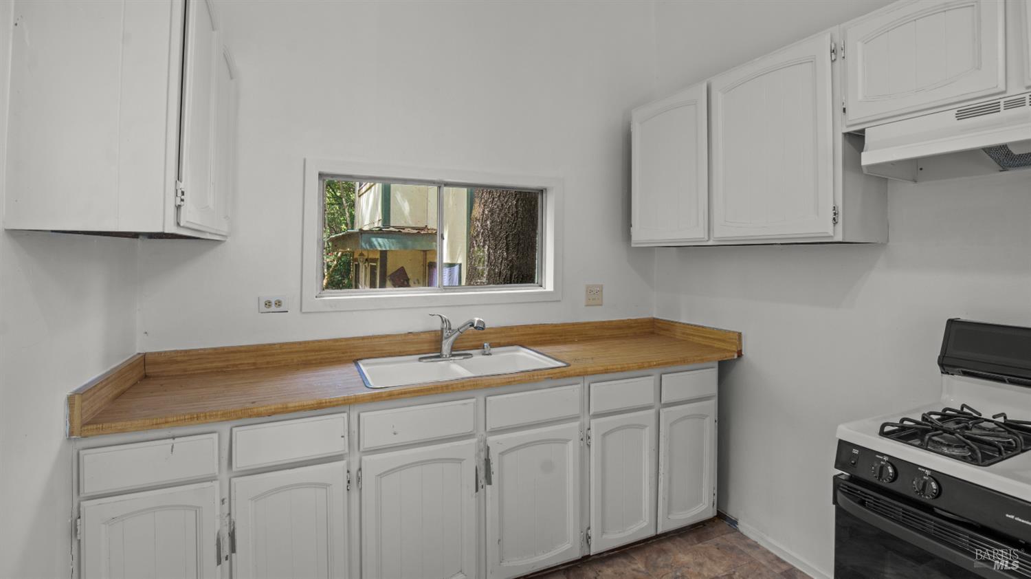 Kitchen with white cabinets, wood-toned counters, a double sink, and a window overlooking a large tree.