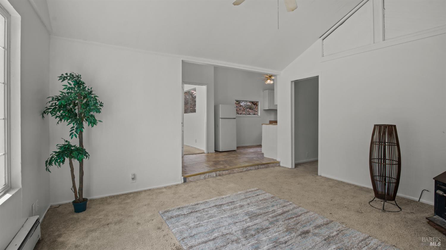 Living room with vaulted ceiling and carpet, opening to a kitchen with a white refrigerator.
