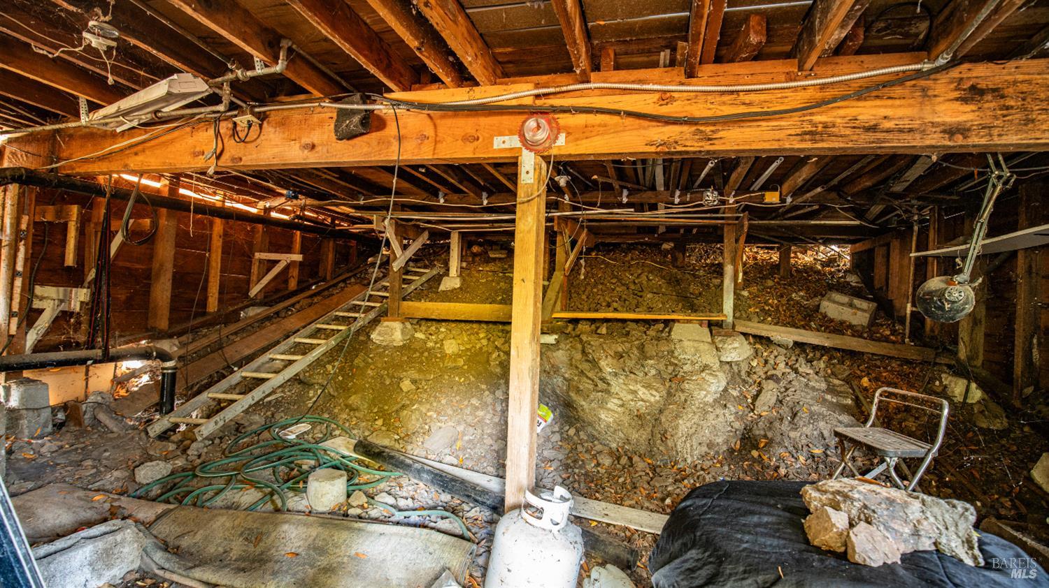 Home crawl space with exposed wooden beams, a ladder, and rubble-covered ground.